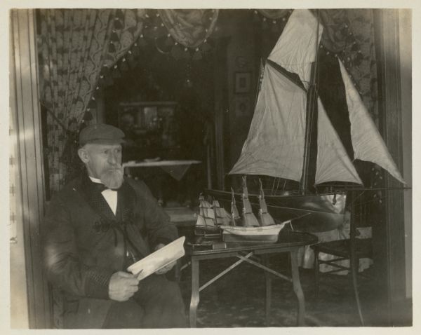 Edward Petersen at home, wearing a smoking jacket with quilted collar and cuffs. He is sitting in a wide doorway, hanging with tasseled draperies, holding a carving knife and a wooden model boat hull. In the room behind him are three models of sailing ships; there are two small models on a low table, and a very large model behind them.