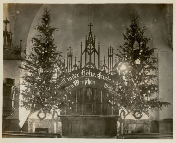 Interior view of a church with large Christmas trees on either side of the Neo-Gothic altar. Between the trees is an arched sign proclaiming, in German, "Glory to God in the Highest, Peace and Good Will to Men." The trees are lavishly decorated with glass ornaments, garlands, and lily flowers, and many candles. On the reverse of the photograph is written: "Leitz Church."