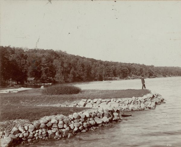 View across water towards Frank Greene standing on a boulder seawall at Uihlein's Point on Geneva Lake. In the background on the left is a pier with a covered pavilion near the shore, and a stairway leading up the hill. A stream enters the lake through a gap in two portions of the wall. This area was part of the Edward Uihlein estate, Forest Glen.