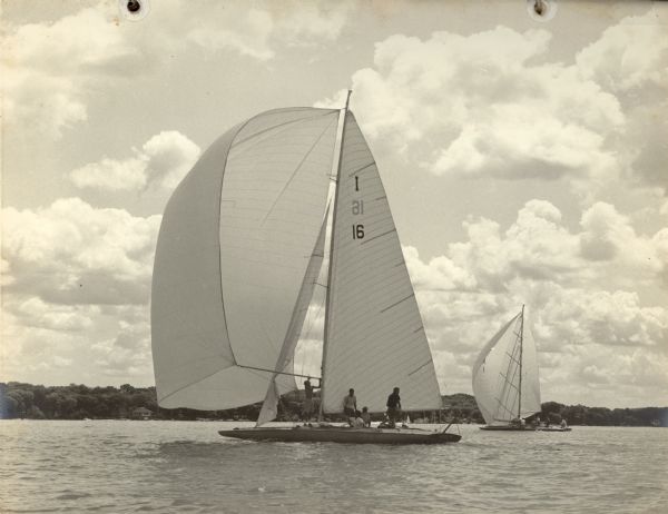 View across water of the <i>Senta III</i>, owned by Ernst Schmidt of Black Point Estate, competes in the Class A Regatta at Pewaukee Lake. The skipper is Buddy Melges. There is a second yacht to the right. Both boats are flying symmetric spinnakers; one crewmember on the <i>Senta III</i> is working with the spinnaker pole. There are large cumulus clouds in the sky.