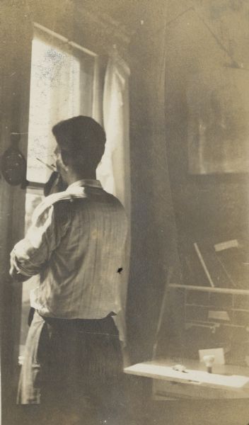 Ernst Schmidt shaving using a straight razor in front of a small mirror. The mirror hangs on the wall next to a window. His shaving brush rests on a drop front desk on the right.
