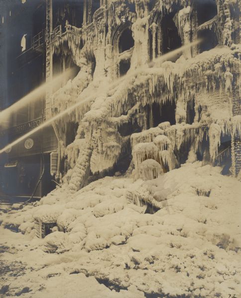 Streams of water from fire hoses are trained into the upper windows of a storefront which is encased in thick ice. A written description of this photograph identifies the building as "Alfred Peats & Company wall paper store on the East side of Wabash Avenue between Madison & Monroe Sts. attacked by fire at six-fifteen in the evening on January 28, 1918." The building on the right was the Chicago office of the Yawman & Erbe Manufacturing Company; the home office was in Rochester, New York.