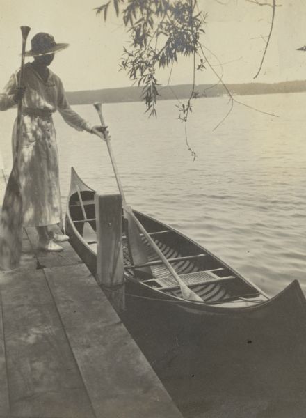 Catherine (Kate) Bartholomay (1899-1991) stands in dappled shade on the pier at Black Point. She is holding two oars as she prepares to get into a wooden canoe that is tied to the pier. Catherine was a granddaughter of Chicago brewer Conrad Seipp, who built Black Point in 1888.