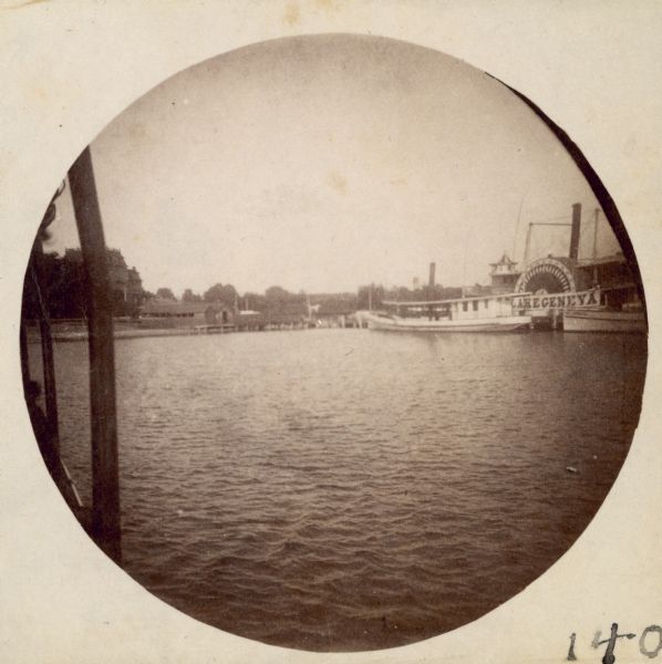 View from boat towards the waterfront at Lake Geneva with the Whiting House Hotel on the left and the pier extending to the right. A large sidewheel steamboat with a banner reading "Lake Geneva" and two smaller launches are on the right.