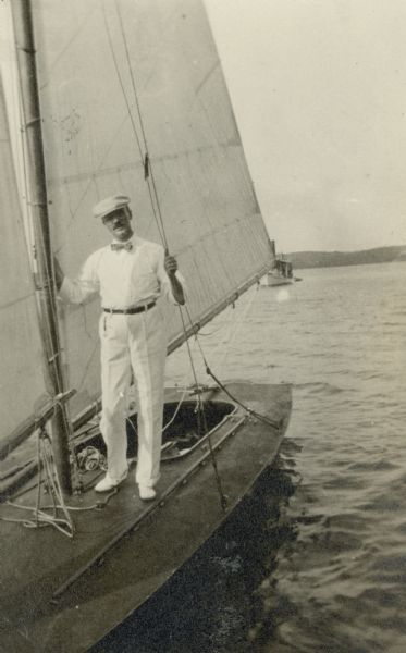 Albert Fridolin Madlener (1868-1947) poses on the small sailboat "Water Witch" at the pier at Black Point. He is wearing a cap, summer slacks, a long-sleeved shirt, and bow tie. There is a larger launch in the background.