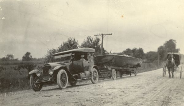 A National brand automobile is stopped on the side of an unpaved road as a horse-drawn buggy approaches from the rear. The car is pulling a racing yacht on a trailer. There are two suitcases on the running board. A description of the photograph written by Ernst Schmidt reads, "Probably coming back from the Oshkosh Regatta in 1920. The old National had a terrible time to tow the boat and the trailer was not successful as it was not heavy enough."