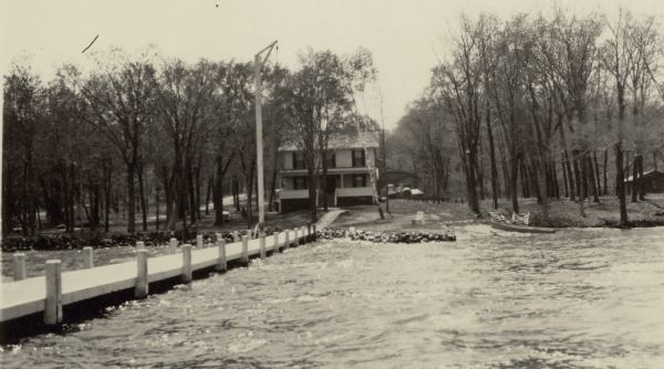 The Lake Geneva Yacht Club, originally a residence, sits on the shore of Geneva Lake. A description of the photograph written by Ernst Schmidt reads, "The picture was taken from the outer end of the pier, looking towards the shore. In those days there was just the main dock. The small pier to the west is still laying on the shore." On the right, tracks lead from the boat ramp onto shore; there is a row boat moored at the ramp.