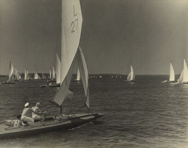 The skipper and crew sail their "E" class boat in a race at the 1933 Inland Lake Yachting Association Regatta on Lake Mendota. Lake View Sanatorium is on the wooded shoreline in the far background.