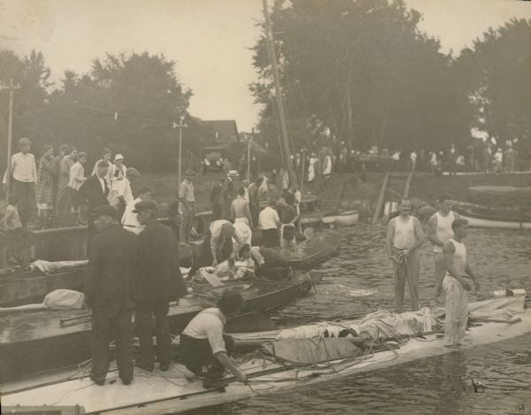 Bystanders watch as men bail out a sailboat and others assess the damage to their craft after a storm hit during the Inland Lake Yachting Association Regatta on Lake Winnebago at Oshkosh. The boats had to be towed to shore because of the damage caused by high wind. There is an automobile on shore in the background.