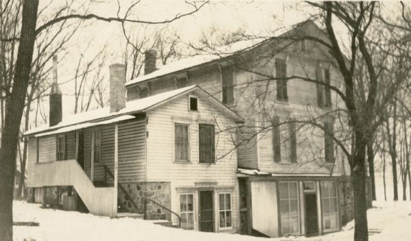 A side and rear view of the Cook house overlooking Geneva Lake. The main structure is a two-story frame building with an exposed basement.  There is a small one-story building, possibly the original house, also with an exposed basement, attached to the rear. Both buildings have stone foundations. There are shutters on the windows of the two-story portion.