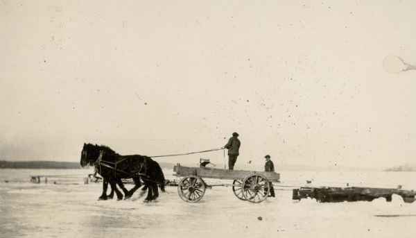 A team of two draft horses, driven by a man standing in a wagon, pulls a portion of crib across the ice of Geneva Lake. Another man walks alongside the wagon. The crib was part of the heavy timber framework used to support the pier at the new club house of the Lake Geneva Yacht Club. Another pier, frozen in the ice, is in the background.