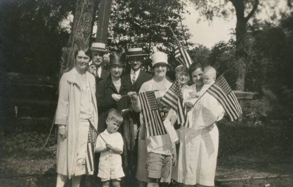 A family group poses on the occasion of the launching of the yacht "Mystery" at Geneva Lake. They are, from left, Alma Schmidt Petersen; Alfred Fridolin Madlener; Conrad Petersen in front of his grandmother, Emma Seipp Schmidt; Dr. Reuter; Elsa Seipp Madlener with Edward Petersen in front; Mrs. Madlener (Alfred's mother), partially obscured; and an unidentified nurse holding the infant William O. Petersen.  Several members of the party wave forty-eight star United States flags. The yacht and its rigging are discernible in the background. Emma and Elsa were daughters of Conrad Seipp, Chicago brewer and founder of Black Point Estate. William O. Petersen donated the estate to the State of Wisconsin in 2005.