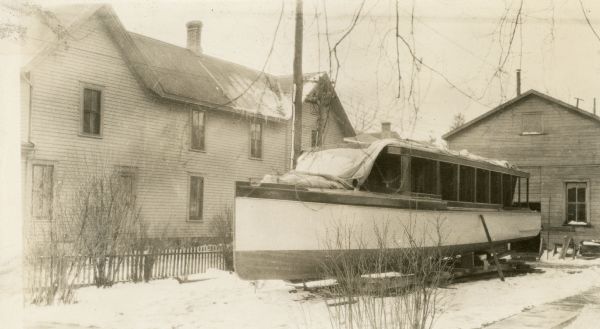 Covers have been pulled aside to reveal the new motor boat which sits in front of a large shed in Till Stuyvesant's yard in Lake Geneva. There is a large two-story frame house on the left. Snow is on the ground. The boat would later be Christened "Nepenthe" and served the residents of Black Point.