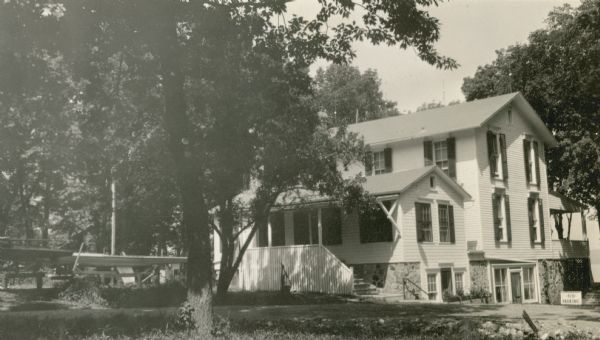 A rear view of the clubhouse of the Lake Geneva Yacht Club. Formerly the Cook house, the building was remodeled in 1927 to accommodate the club. The building has been repainted and there are new shutters on the windows. A sailboat is in the yard to the left. There is a glimpse of Geneva Lake on the far right.