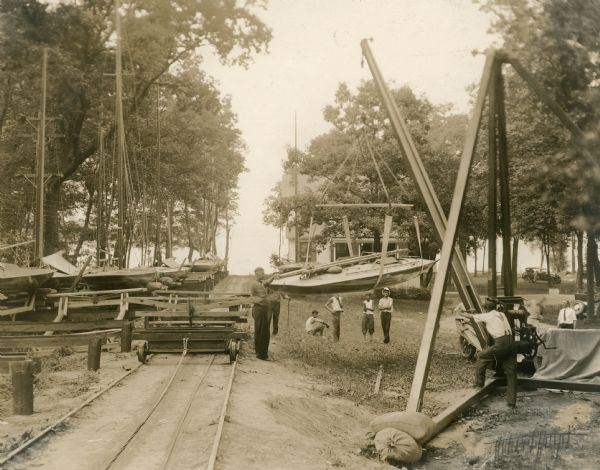 Men use a derrick to lift a racing yacht from its trailer, obscured by the derrick's winch, onto the car on the tracks leading to Geneva Lake. The boat was competing in the Inland Lake Yachting Association Regatta. The Lake Geneva Yacht Club clubhouse is in the background. There are several sailboats on racks on the left.