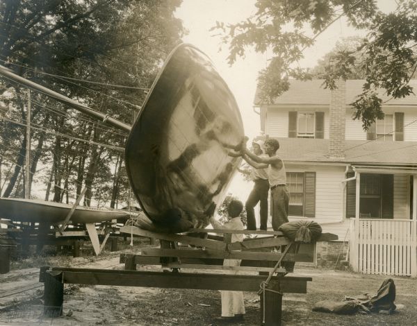 Members of the crew work on the bottom of a class C boat before the Inland Lake Yachting Association Regatta. The Lake Geneva Yacht Club clubhouse is in the background. On the left are the tracks leading to the boat launch; there is another sailboat on a rack on the left.