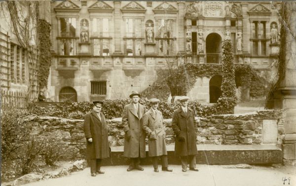 Ernst Schmidt, second from left, on a tour of the ruins of Heidelberg Castle with his guide, left, and two unidentified persons. They are standing in the courtyard in front of the facade of the ruins of the sixteenth century palace of Elector Otto Henreich. There is open space behind the upper portion of the wall behind the group. There are statues in niches between the windows.