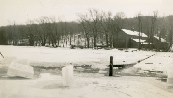 View of the Black Point boathouses taken from the frozen ice of Geneva Lake. Blocks of cut ice are in the foreground. A description of the photograph written by Ernst Schmidt reads, "The channel cut, after the weight for the "Nepenthe" had been dropped to the bottom on February 11. The hole was 220 feet from shore and in 76 feet of water." A sailboat is stored on a rack on a section of pier to the left of the boathouses.