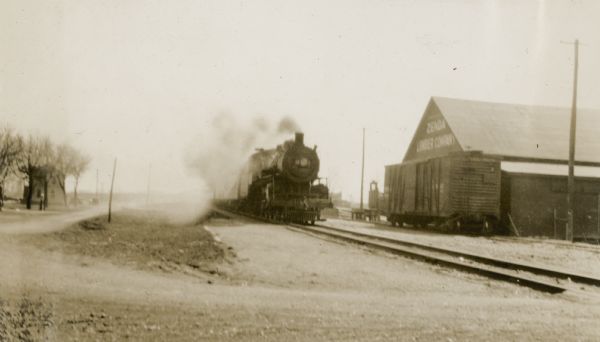 Ernst Schmidt has captioned this photograph, "The C. M. & St. P. train arriving at Zenda at 10:30 A. M. on Sunday, April 12. Alma, Edward and Conrad came up on that train." The steam engine is approaching the Zenda Lumber Company; there is a freight car on the siding. An unpaved road crosses the tracks and another runs alongside the tracks. There is a house in the background on the left.