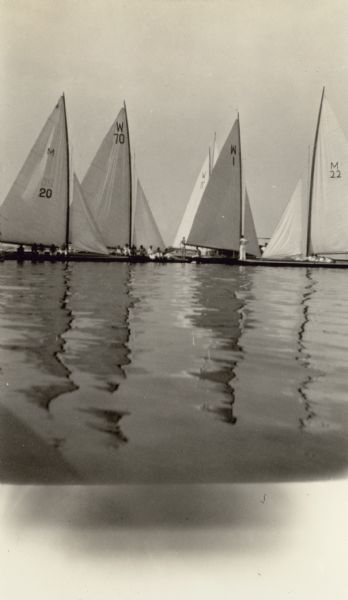 View across water towards sailboats with their crews reflected in the still waters of Lake Winnebago, before the start of a race at the Inland Lake Yachting Association Regatta at Oshkosh.