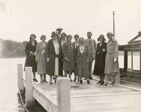 Albert Madlener, in rear with cap, and Ernst Schmidt, third from right, pose with a group of nine women officials of Chicago's Grant Hospital. In a written description, Ernst Schmidt has identified them, from left, as Miss Mikkolo, Miss Olson, Miss Watson, unidentified, Miss Larson, Miss Sheer, Miss Schock, unidentified, and Miss Tegge. A launch is moored to Madlener's dock. The group was about to depart on a boat ride.