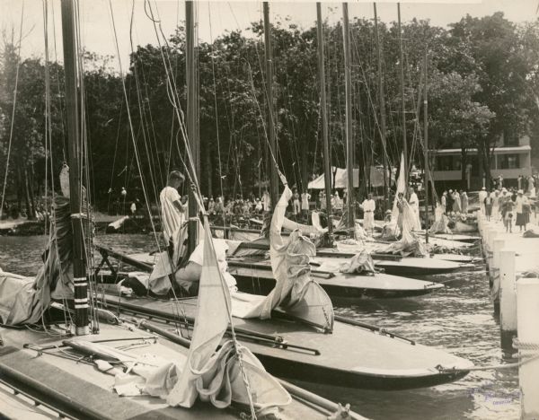 Crews work on their boats at the dock of the Lake Geneva Yacht Club after one of the races of the 1932 Inland Lake Yachting Association Regatta. The tent for spectators is in the background on the shoreline to the left of the clubhouse. People are gathered along the shore and also strolling on the pier.
