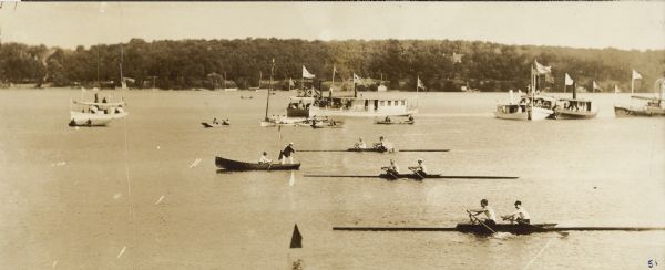 Slightly elevated view across water towards the starting line of a two-oared scull race on Geneva Lake. At the front of the line a judge wearing a dark jacket and light pants stands in a rowboat with another man at the oars. There are many spectators in the background in steam launches and row boats. Flags fly from the larger craft.