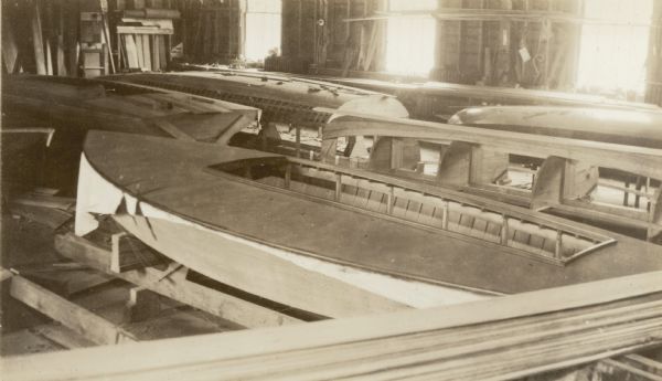 Several 16 foot sloops are shown at various stages of construction in a building at the Geneva Lake Boat Company. Untrimmed canvas extends from the deck of the boat in the foreground.