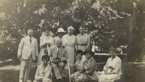 Group portrait of eight women and two men posing on the lawn at Black Point. The description on the reverse of the photograph reads: "The Wacker family after [they] came for Sunday dinner." The individuals are identified as, standing left to right: Mr. Harry Williams, Mrs. Elsa (Seipp) Madlener, Mrs. O.L. (Emma Seipp) Schmidt, Mrs. Conrad (Catherine Orb) Seipp, Mrs. August Magnus; center, seated in chair, Mr. Charles Wacker; seated on ground left to right: Rosalie Wacker, Mrs. Frederick Wacker, Mrs. Junkin, Mrs. Harry Williams.