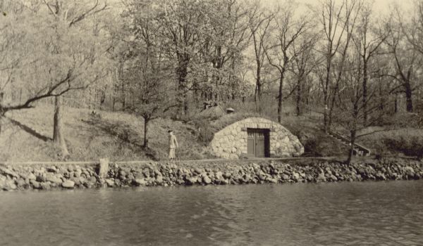 View across water towards Emma Seipp Schmidt walking along the shore path at the estate of Albert Madlener on Geneva Lake. There is stone riprap supporting the bank. On the right is a stone-faced boathouse with rooftop ventilators built into a small ravine.
