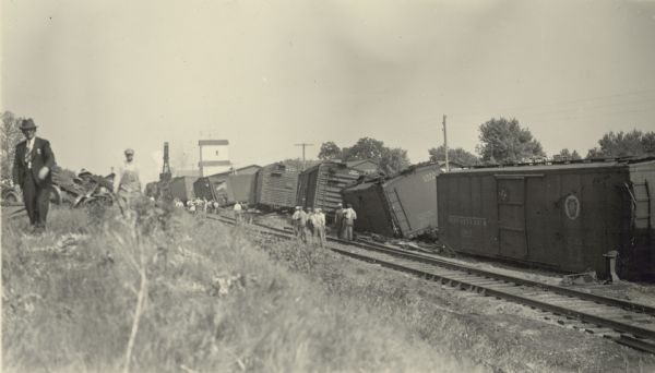 Slightly elevated view from bank of workmen gathered at the site of a train derailment. The man in suit and hat at far left is identified as a police officer. Damaged undercarriages are behind him. In the background is a grain elevator; to the left of the elevator is a derrick brought in to lift the train cars. The cars are nearly upright despite having lost their undercarriages.