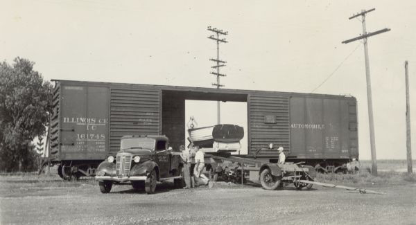 Ernst Schmidt, second from left, and two other men prepare to unload a 17-foot Lyman motorboat named "Islander" from an Illinois Central freight car. "Automobile" is painted on the side of the freight car. A boat trailer is positioned to load the boat and a General Motors truck is parked on the left. The printing on the door of the truck reads, "ERNST C SCHMIDT, BLACK POINT, LAKE GENEVA WIS." A railroad crossing sign is visible through the open doors of the freight car.