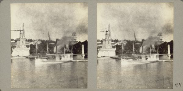 Elevated view across water towards an unidentified man standing on the deck of the steamship "Arrow," owned by Shelton Sturges. It is docked at a pier near a large windmill which was part of the estate of Levi Ziegler Leiter, a Chicago retailer and early partner of Marshall Field. There is a sailboat docked on the right. Other buildings are in the background on the hill above the shoreline.