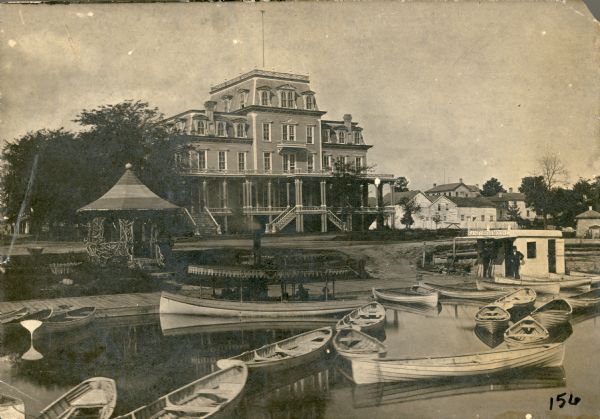 View from water over boats towards the four-story wood frame Second Empire style Whiting House Hotel standing on the lake shore at Lake Geneva. Several sets of stairs lead to a porch which extends across the front of the first floor. A rustic gazebo stands at left, and other buildings of the village are seen on the right. A wharf extends along the waterfront and several rowboats, and a small steam launch with passengers, are moored in front of the hotel. Two men stand in front of a small building on the right with the sign "Sanford and Moore" above its awning.