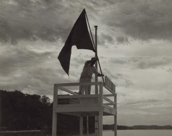 The caption accompanying this photograph describes the scene: "Ernst C. Schmidt raising the storm warning flag at the dock of the Lake Geneva Yacht Club on Friday, July 25, 1952. This picture was taken for the Water Safety Patrol Committee."