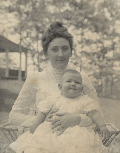 Clara Seipp Bartholomay, a daughter of Chicago brewer Conrad Seipp, sitting outdoors in a wicker chair, holding her infant daughter Catherine Bartholomay on her lap. Clara is wearing a light-colored dress with long sleeves and high collar. There is a porch or other open structure in the background.