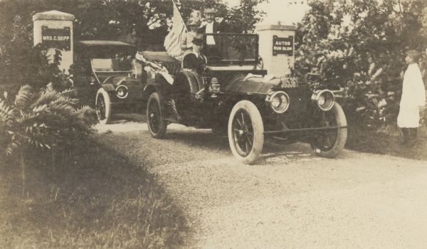 A well-dressed couple stand in the back of an chauffeur-driven open automobile on the drive at Black Point Estate. The woman, who is wearing a very tall hat, holds an American flag. There is a pillar on each side of the drive; the left pillar has a sign that reads: "Mrs. C. Seipp," and on the right pillar, "Autos Run Slow." A boy stands watching on the right.