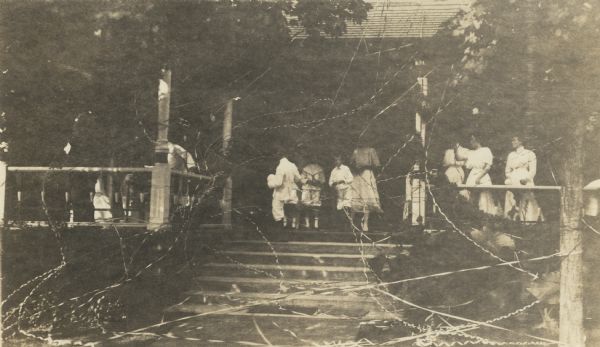View from lawn towards a group of women and children standing on the porch at Black Point. The porch, stairs and trees are strewn with streamers, part of the celebration of the marriage of Alma Seipp, daughter of Conrad Seipp, to William Sherman Hay.