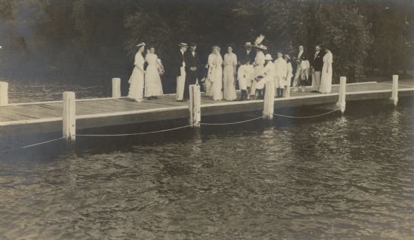 View across water towards a group of well-dressed adults and children gathered on the pier at Black Point Estate on the wedding day of Alma Seipp and William Sherman Hay. Dr. Otto Schmidt, husband of Alma's sister Emma, is fourth from the left. Behind the group is a tree-lined shoreline.