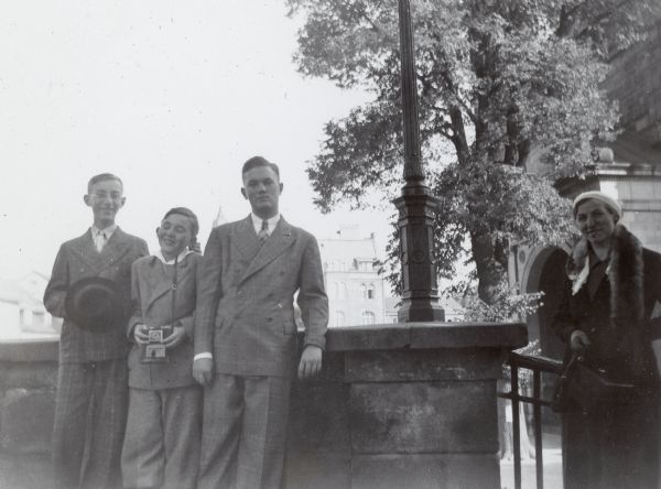 Conrad Petersen, far left, tugs on his younger brother William's ear, while posing with a more serious Edward against a stone wall in Eisenach, Germany. William  is holding a camera. An unidentified woman smiles on the far right. Eisenach is the birthplace of Johann Sebastian Bach; nearby Wartburg Castle provided a refuge for Martin Luther.