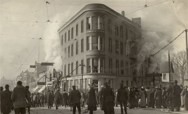 Smoke billowing from windows on the State Street (left) and Carroll Street (right) sides of the Wisconsin Building. Fire fighters on ladders are working from both sides of the building as a crowd is watching. The building housed the Commercial National Bank and Keeley's Palace of Sweet's, known at the time as "The Pal." The dome of Bascom Hall on the University of Wisconsin campus is at the end of State Street on the far left. Along the street are streetcar tracks and overhead lines. A garland of evergreens are hanging from wires strung on utility poles along the sidewalk.