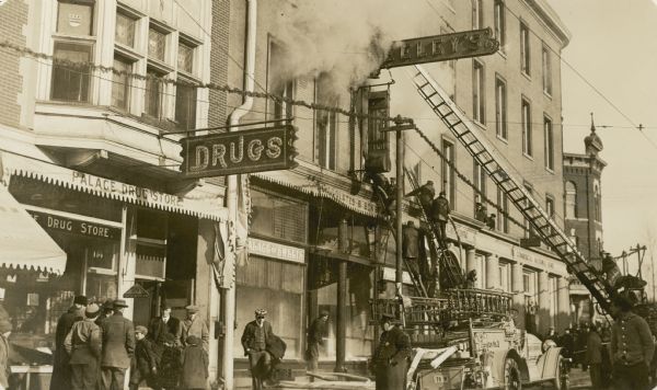 A policeman, center, keeping an eye on bystanders as firemen on ladders fight a fire at the Wisconsin Building on State Street. The building housed the Commercial National Bank and Keeley's Palace of Sweets, also known as "The Pal." A garland of evergreens is hanging from wires strung along utility poles above the sidewalk. Several men are leaning out of a second story window on the right.