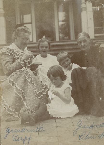 Mrs. Conrad (Catherine Orb) Seipp, left, and Mrs. Ernst (Theresa Weikhardt) Schmidt join their grandchildren, from left, Alma Catherine, Clara Theresa (Tessa), and Ernst Conrad Schmidt for tea in front of the porch steps at Black Point. Mrs. Seipp is wearing a light-colored dress with ruffles; Mrs. Schmidt is in a plain dark dress.