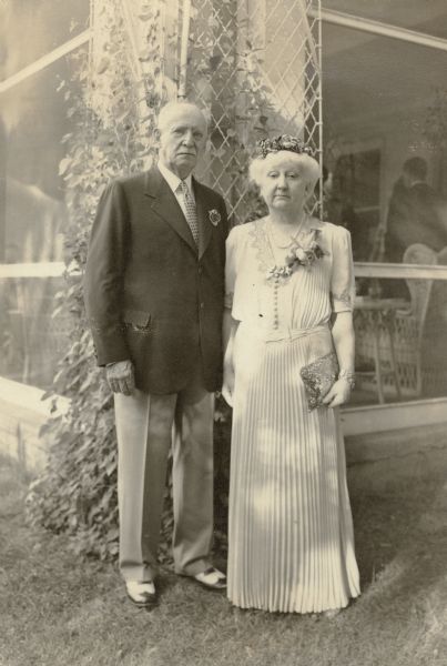 Henry Bartholomay, Jr. and his wife, Clara Seipp Bartholomay pose at the corner of the screened porch at their summer home on the occasion of their fiftieth wedding anniversary. The house was built by Clara's mother, Mrs. Conrad Seipp in 1905 on the grounds of Black Point Estate. There is a trellis with vines behind them. A wicker chair and table, as well as guests, are visible through the screen. Clara wears a hat and corsage; Henry has a boutonniere.