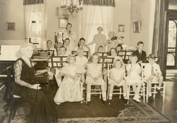 Descendants of Conrad Seipp comprise an orchestra which performed a concert as part of the celebration of the fiftieth anniversary of the construction of the main house at Black Point. Emma Seipp Schmidt sits at the piano in the music room of the house. In the background, Alma Seipp Hay holds her violin. Alma Schmidt Petersen stands to the left of her Aunt Alma Hay. Alma Petersen's sons, Conrad, seated in back row at extreme left, Edward, standing with toy horn, and William, second row, far right, were in the orchestra.