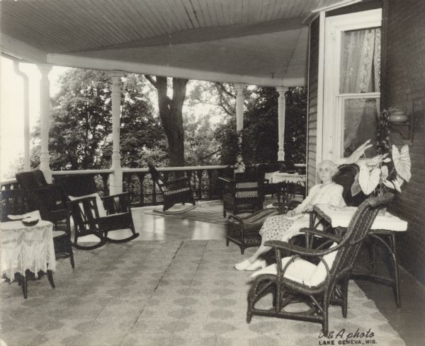 Alma Schmidt Petersen poses seated in a wicker chair on the porch at Black Point. She is wearing a dress and light coat. There is a large-leafed potted plant on a table beside her, and other wicker furniture is arranged on the porch. There are woven seagrass rugs on the floor.
