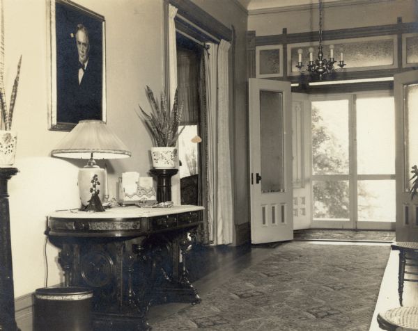 A portrait of Dr. Otto L. Schmidt hangs in the front hall of Black Point. A handwritten caption which accompanies the photograph states: "Portrait above — Otto L. Schmidt by Klemperer -—1929." A library table with lamp stands below the portrait; beside it is a potted plant on a tall stand. The double entry doors are open onto the porch.  An interior doorway with draperies is to the left of the entrance.