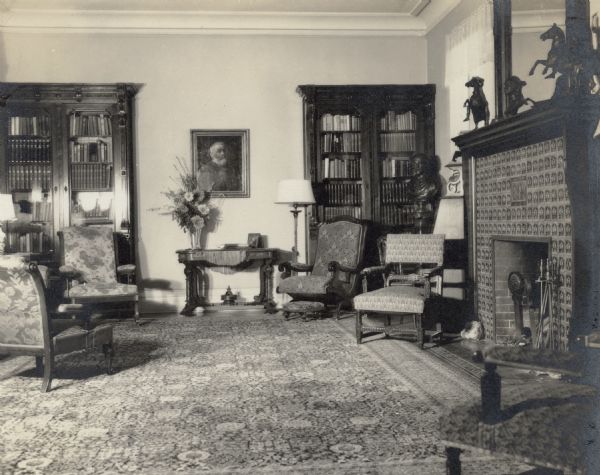 Two tall bookcases stand against the far wall in the living room at Black Point. A portrait of Conrad Seipp hangs on that wall. A large bouquet in a tall vase stands on a side table below the portrait. On the right is a fireplace with a tile surround. There are two bronze horses on the mantle.