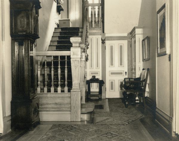 A grandfather clock stands to the left of the stairway in the front hall of Black Point. There is a paneled door in the background and a settee on the right.