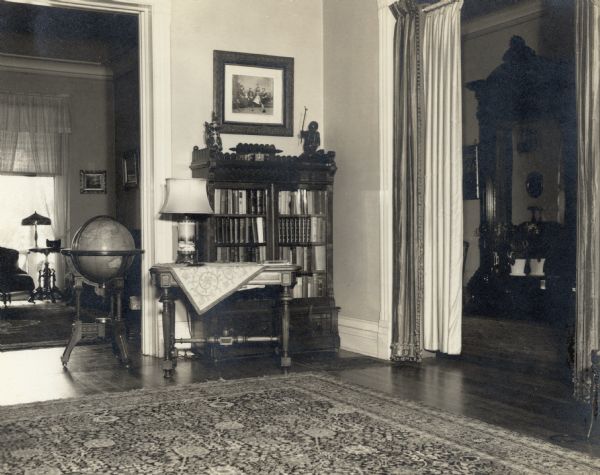 A view of a corner of the living room at Black Point provides a glimpse into the music room, at left, and the front hall, right. In the living room is a bookcase in front of the far wall, and a side table in front of it. A floor globe stands in the doorway. There is a large pier mirror in the hall.